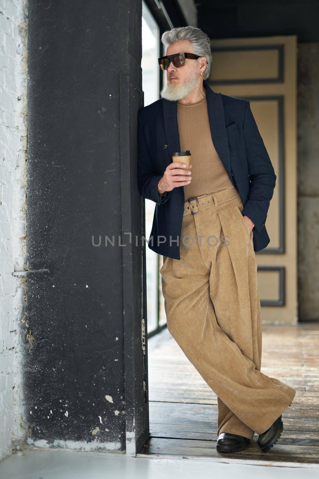 Full length shot of stylish middle aged man in business casual wear and sunglasses leaning on the wall, holding disposable coffee cup while posing in loft interior by friendsstock