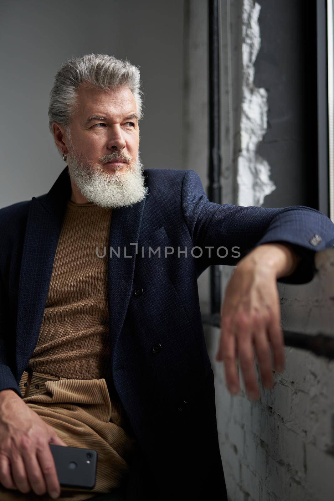 Handsome stylish mature man in business casual wear looking out a window, holding smartphone while sitting in modern loft interior by friendsstock