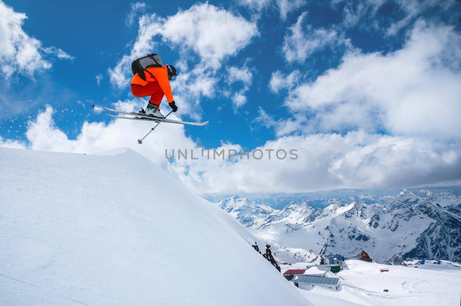 A professional skier in an orange suit jumps from a high cliff against a background of blue sky and clouds, leaving a trail of snow powder in the mountains