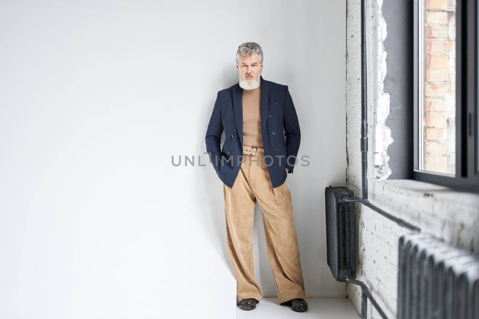 Full length shot of stylish middle aged man in business casual wear looking at camera, putting hands in pockets while posing against white background in loft interior. Lifestyle, people concept