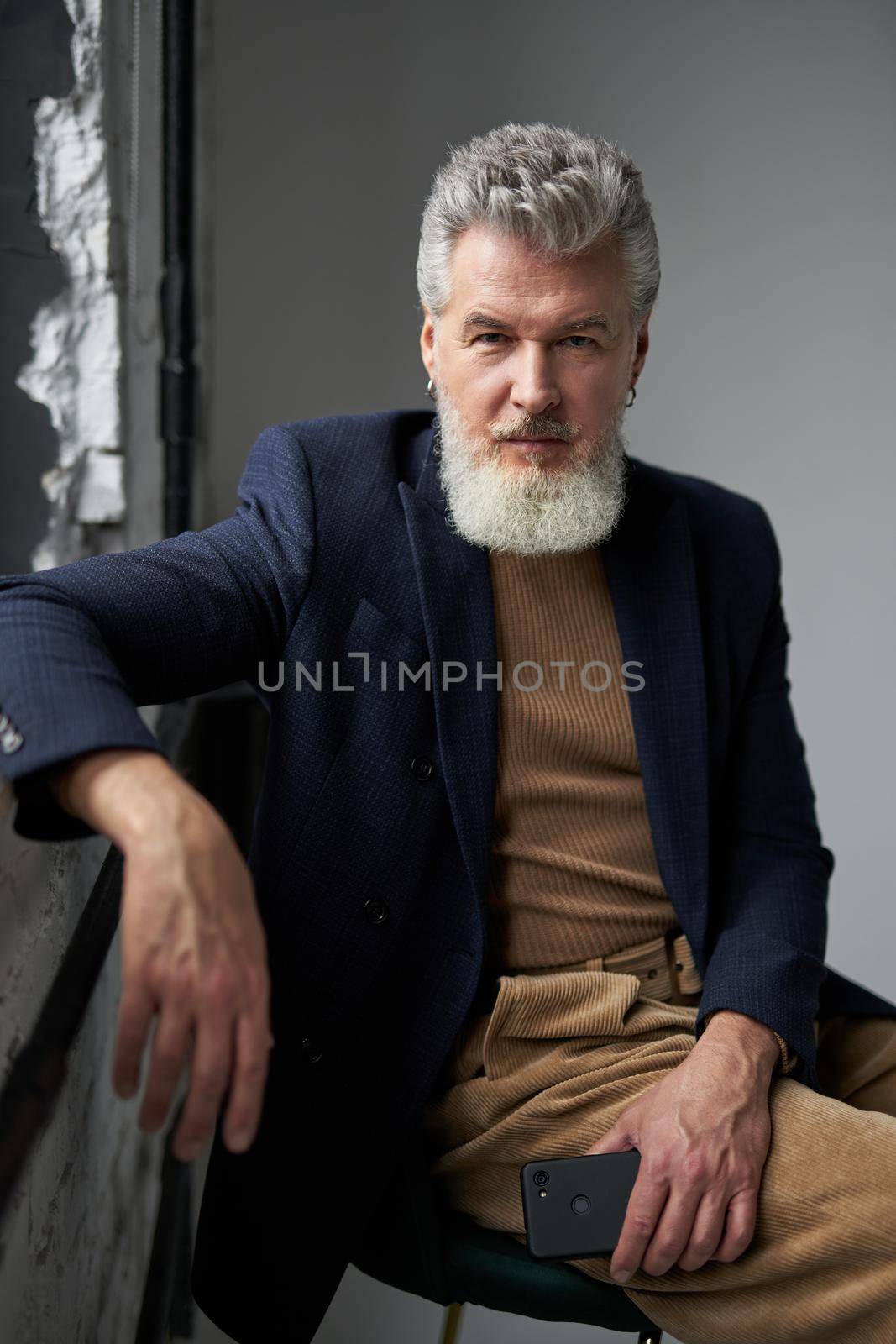 Stylish middle aged man in business casual wear looking at camera, holding smartphone while sitting by a window in modern loft interior. Lifestyle, people concept