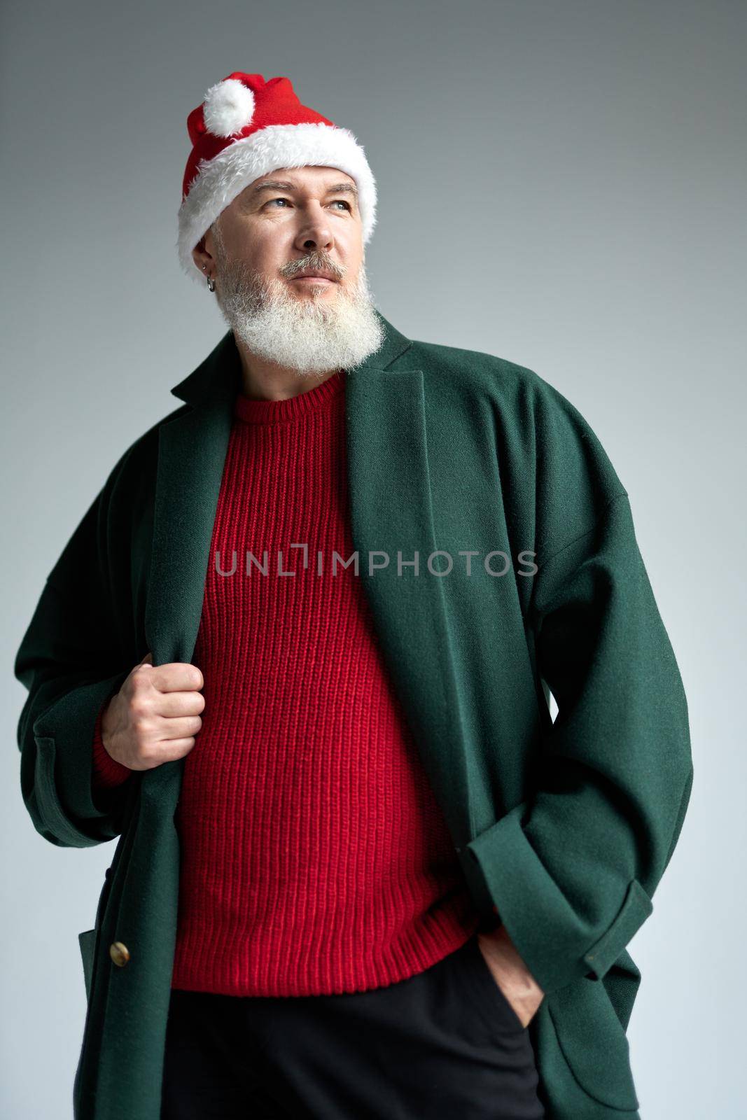Cool middle aged man with Christmas hat wearing stylish outfit looking away, adjusting his coat while posing isolated over light gray background by friendsstock