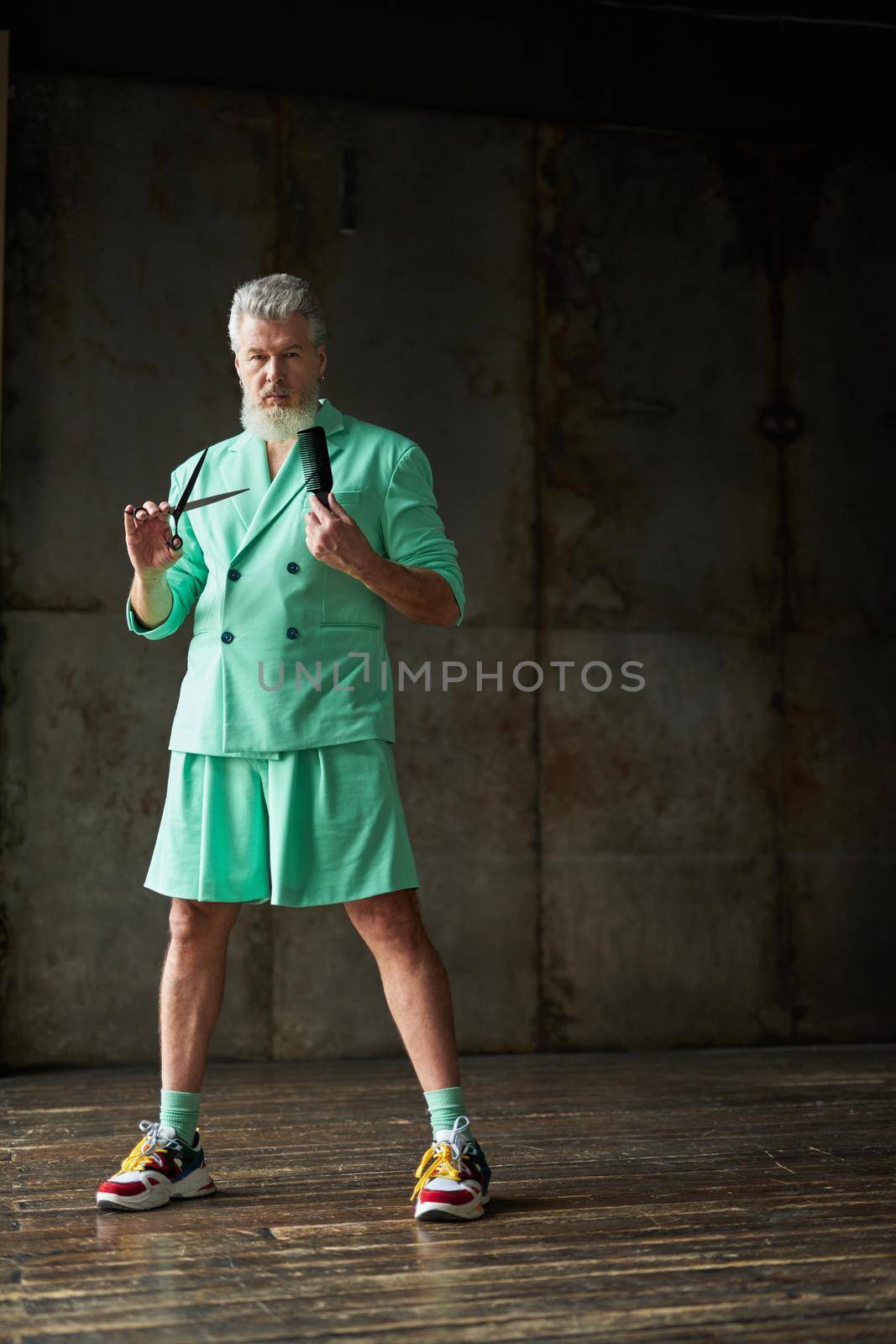 Full length shot of trendy gray haired mature man with beard wearing stylish outfit looking at camera, holding sharp barber scissors and hair comb, standing over dark background by friendsstock