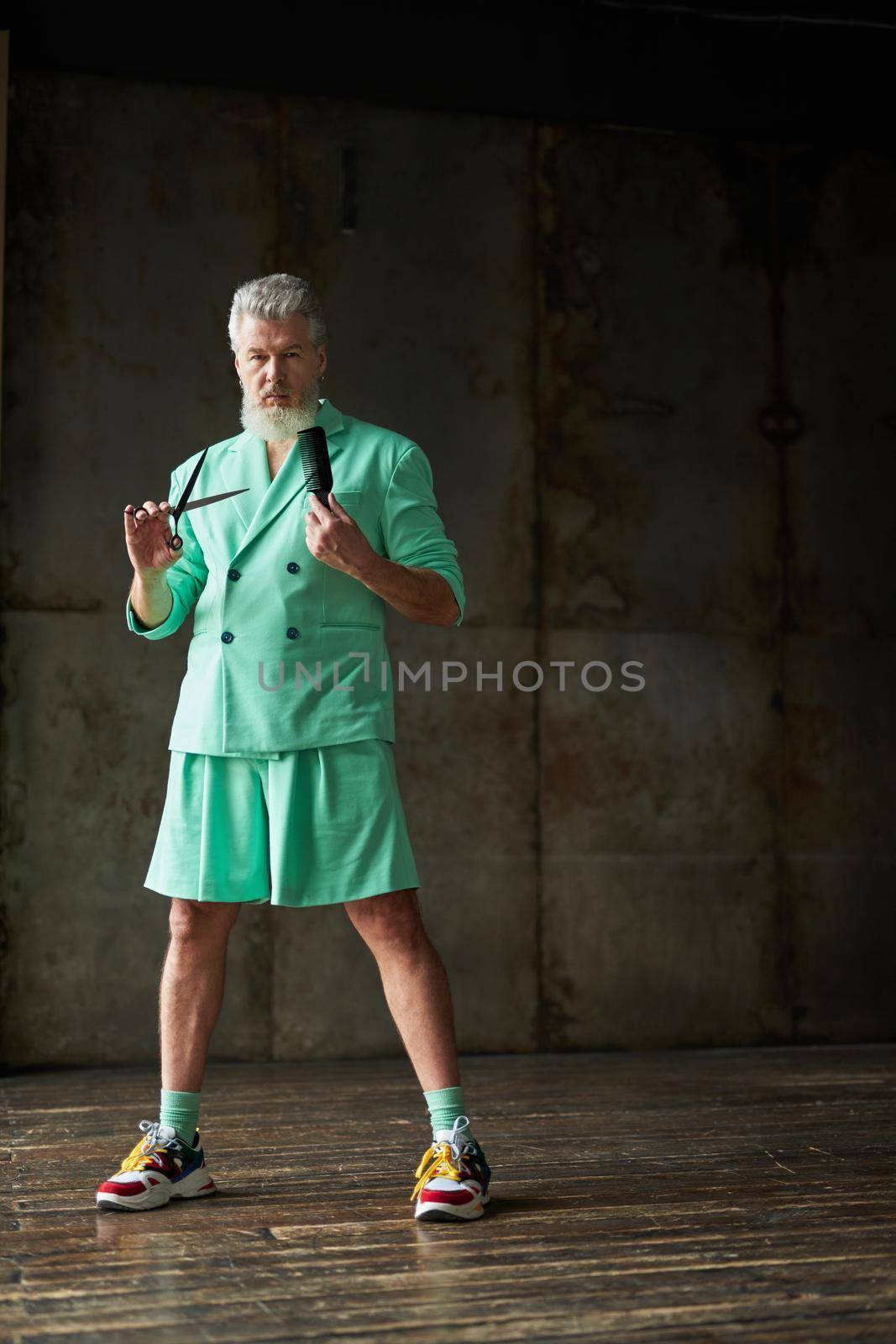 Full length shot of trendy gray haired mature man with beard wearing stylish outfit looking at camera, holding sharp barber scissors and hair comb, standing over dark background