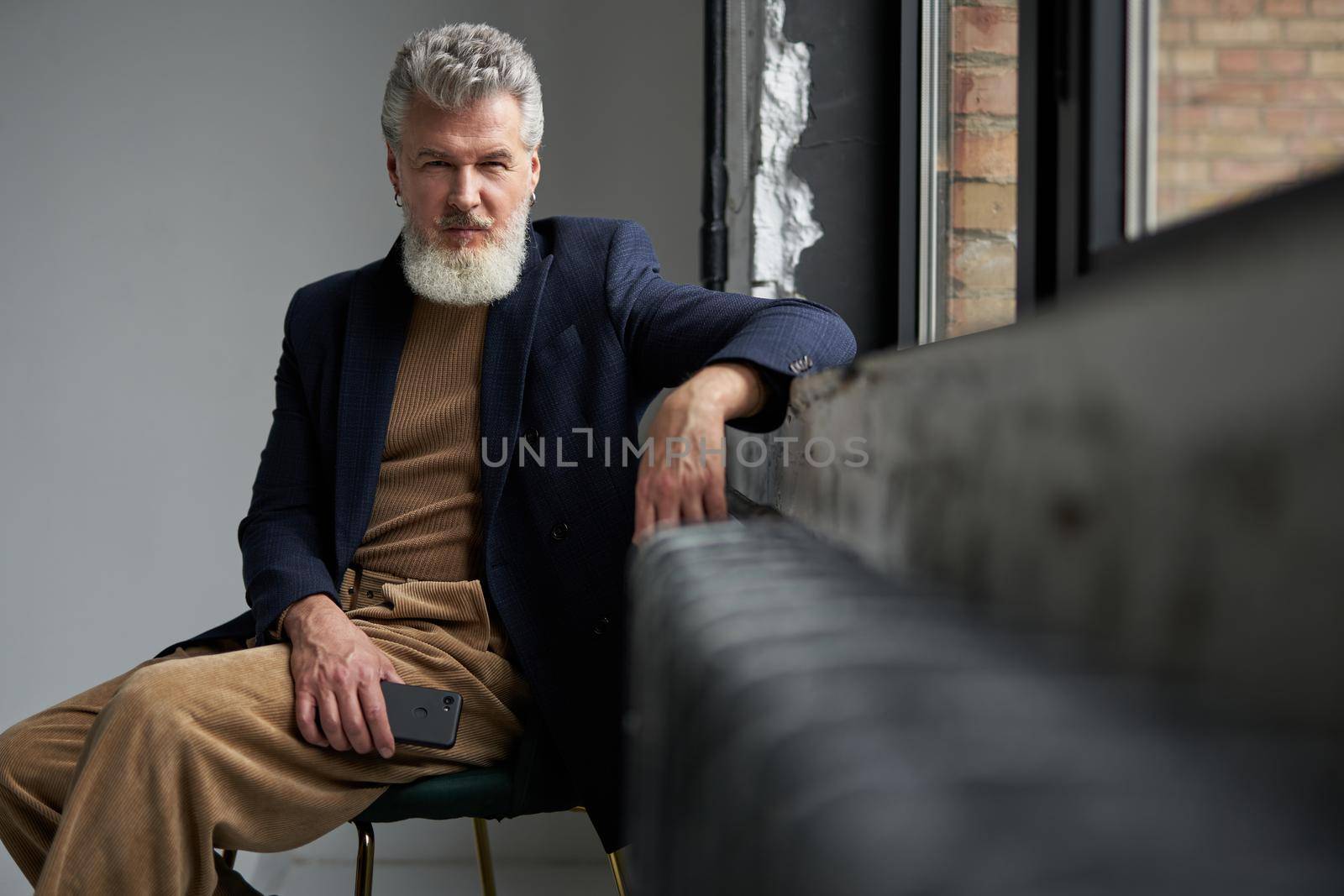 Stylish gray haired middle aged man in casual wear looking at camera, holding smartphone while sitting by a window in modern loft interior. Lifestyle, people concept
