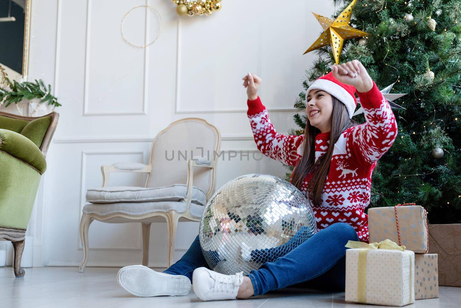 Merry Christmas and Happy New Year. Young brunette woman in santa hat holding mirror disco ball dancing by the Christmas tree