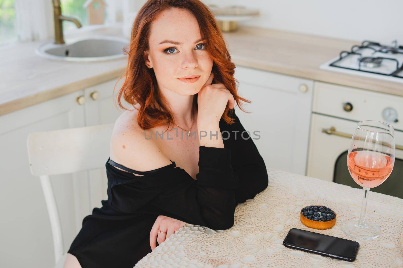 A beautiful girl with red hair holding a smartphone. There's a glass of rose wine on the table. The girl is resting.
