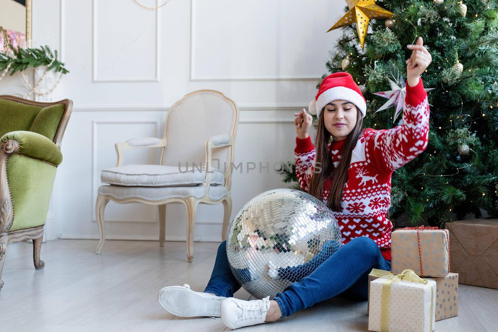 Young woman in santa hat holding mirror disco ball dancing by the Christmas tree by Desperada