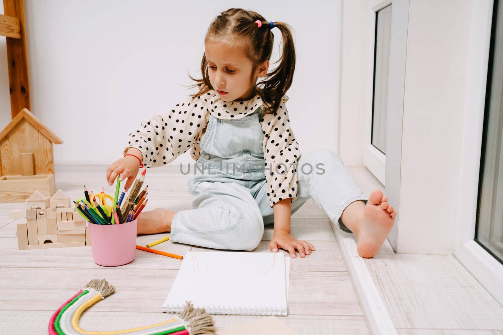A little girl sits on the floor and draws in a sketchbook with colored pencils. The child draws flowers and rainbows. Pencils in the hands of the girl.