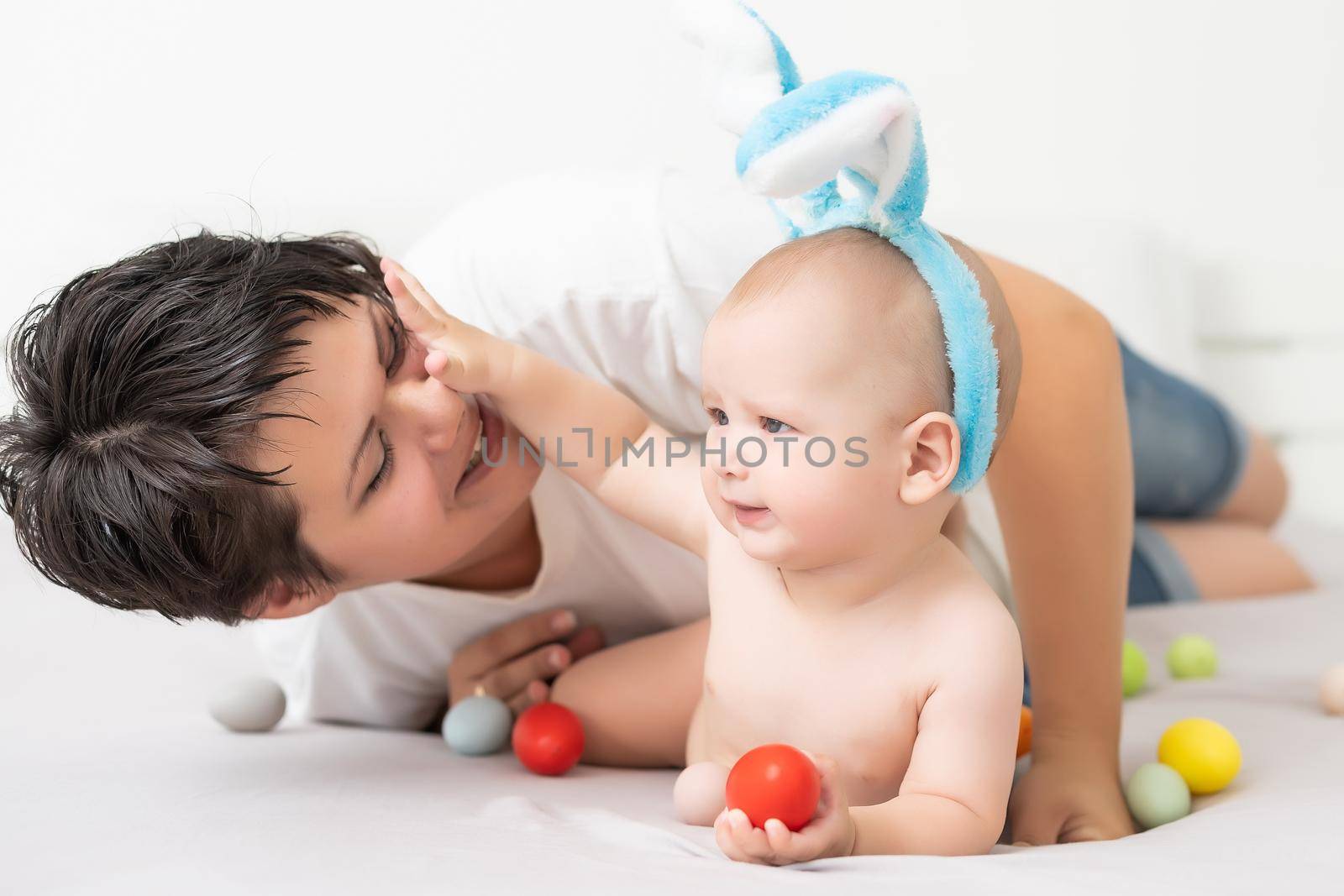 Mom and baby with rabbit ears, with Easter eggs in their hands, sitting on the sofa, parents and children play indoors. Family celebrates Easter by Andelov13