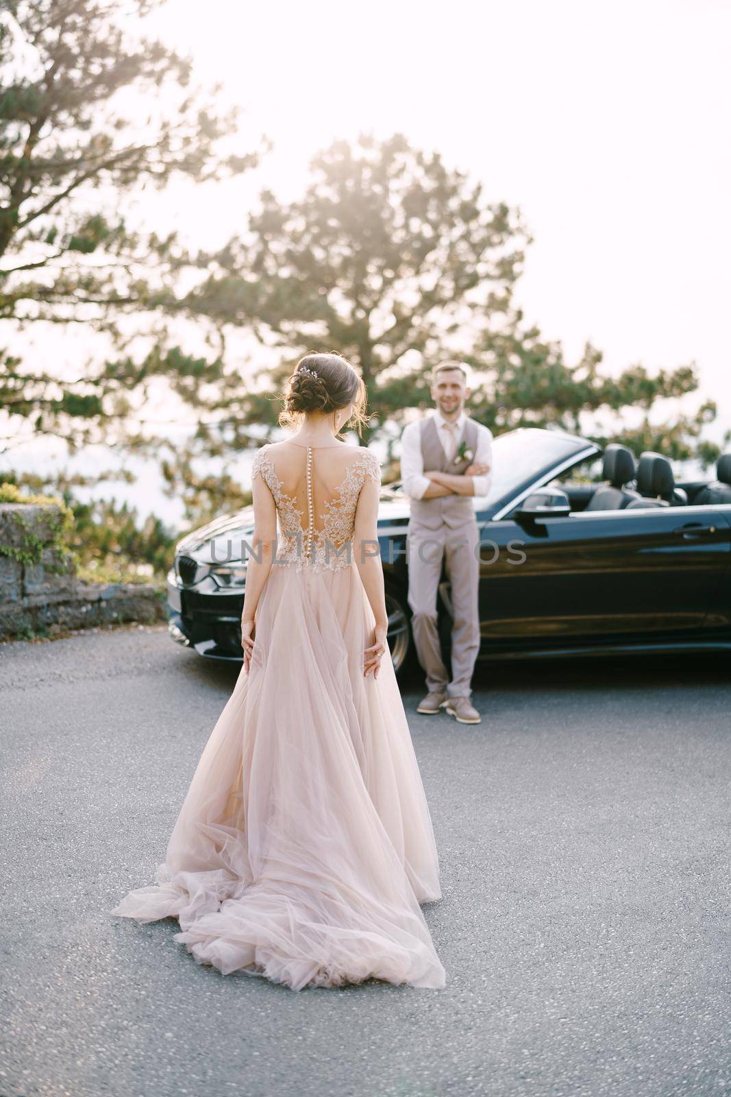 Bride walks along the road to groom standing near the convertible by Nadtochiy