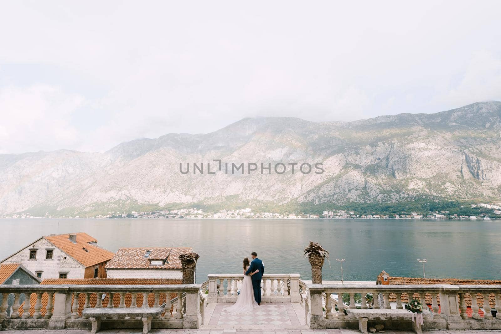 Bride hugs groom on the stone terrace against the backdrop of the bay and mountains by Nadtochiy