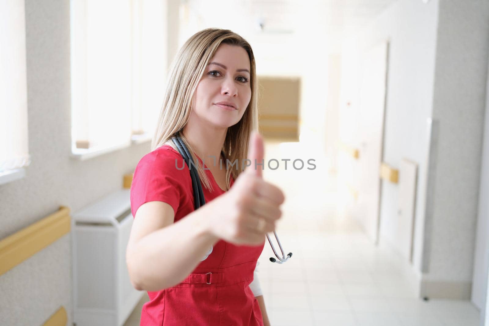 Beautiful female nurse in hospital corridor showing thumbs up, success gesture. Confident hospice worker