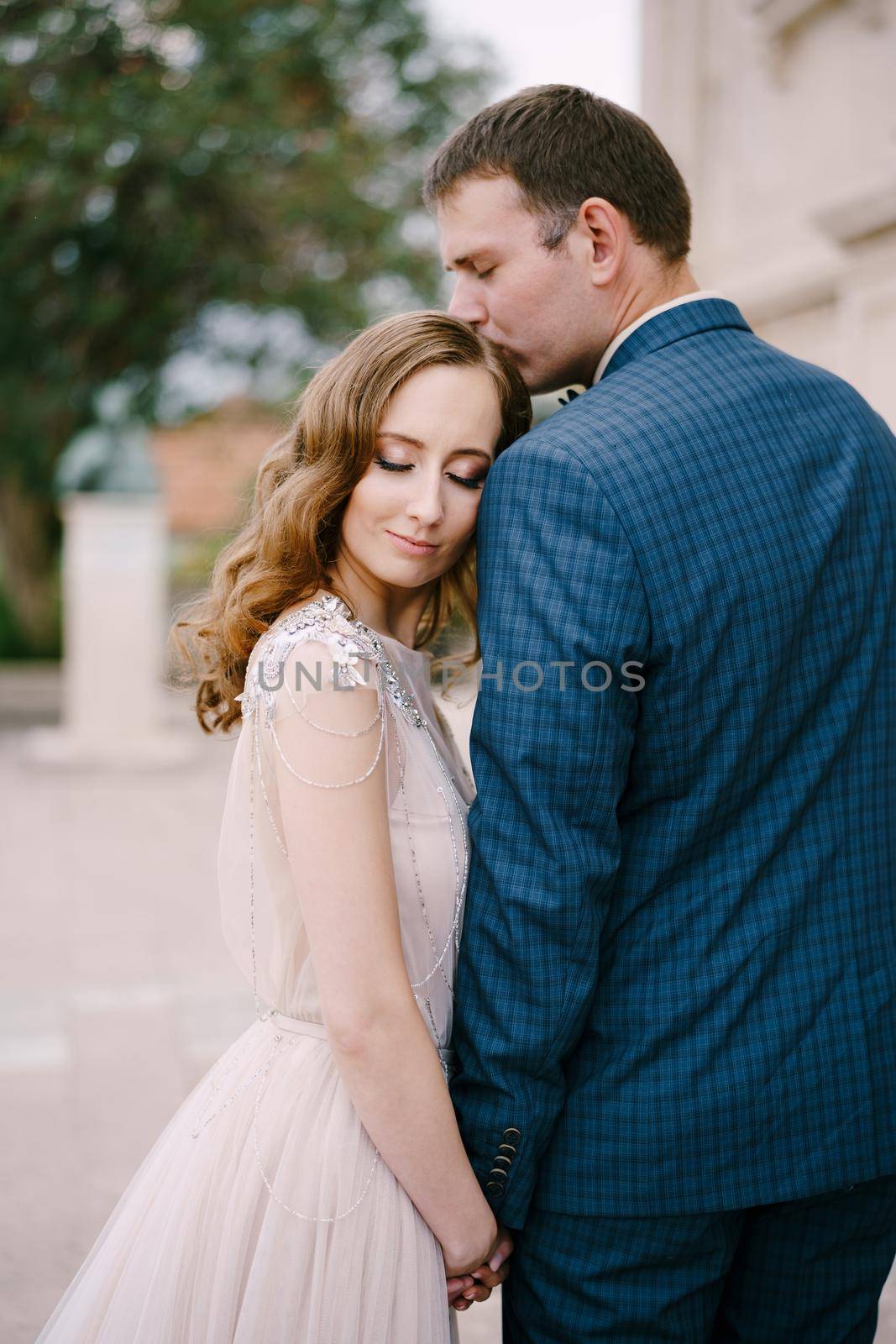 Groom kisses the top of the bride head while holding her hand. Portrait by Nadtochiy