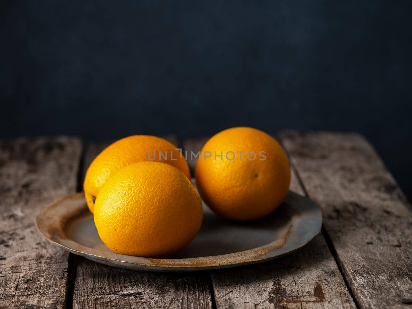 Three oranges on dark background, big dark vintage plate, old wooden table, side view, copy space