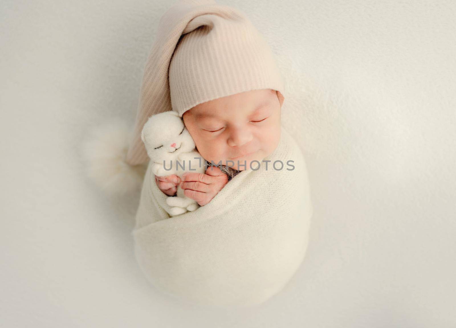 Newborn baby girl swaddled in white fabric holding cute bunny toy and sleeping. Adorable infant child kid napping wearing hat