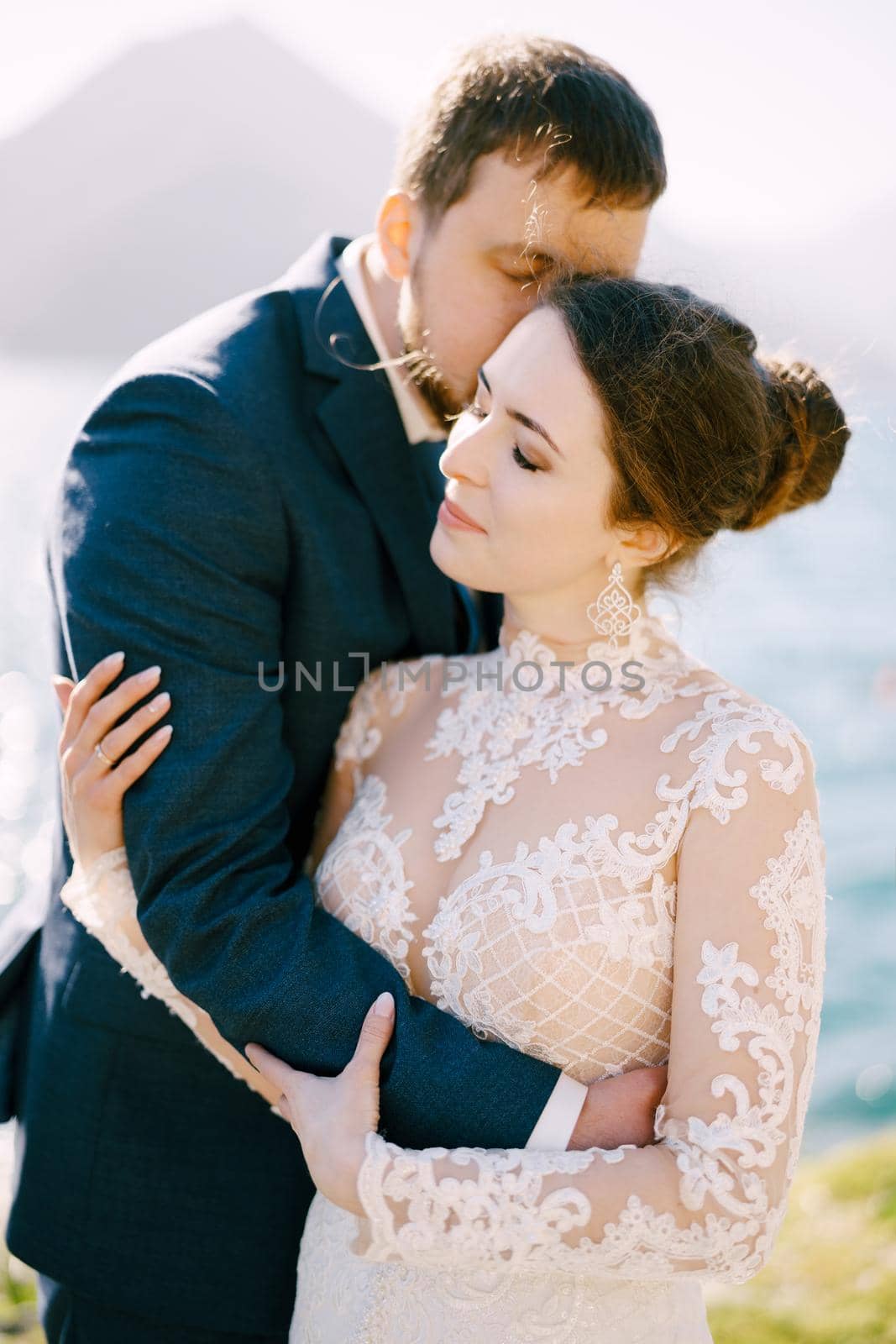 Groom hugs bride and kisses her cheek. Side view. Close-up by Nadtochiy