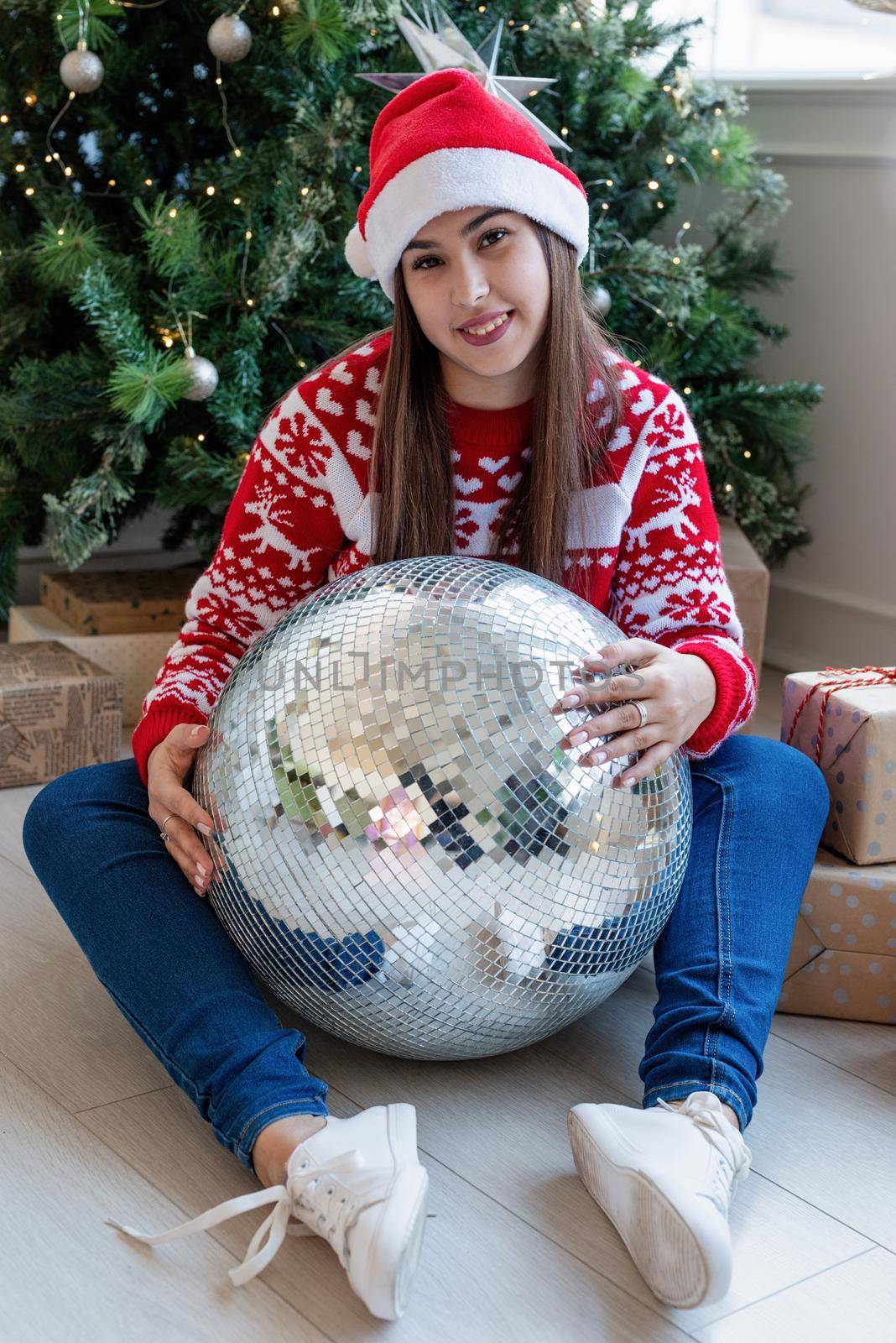 Merry Christmas and Happy New Year. Young brunette woman in santa hat holding mirror disco ball sitting by the Christmas tree