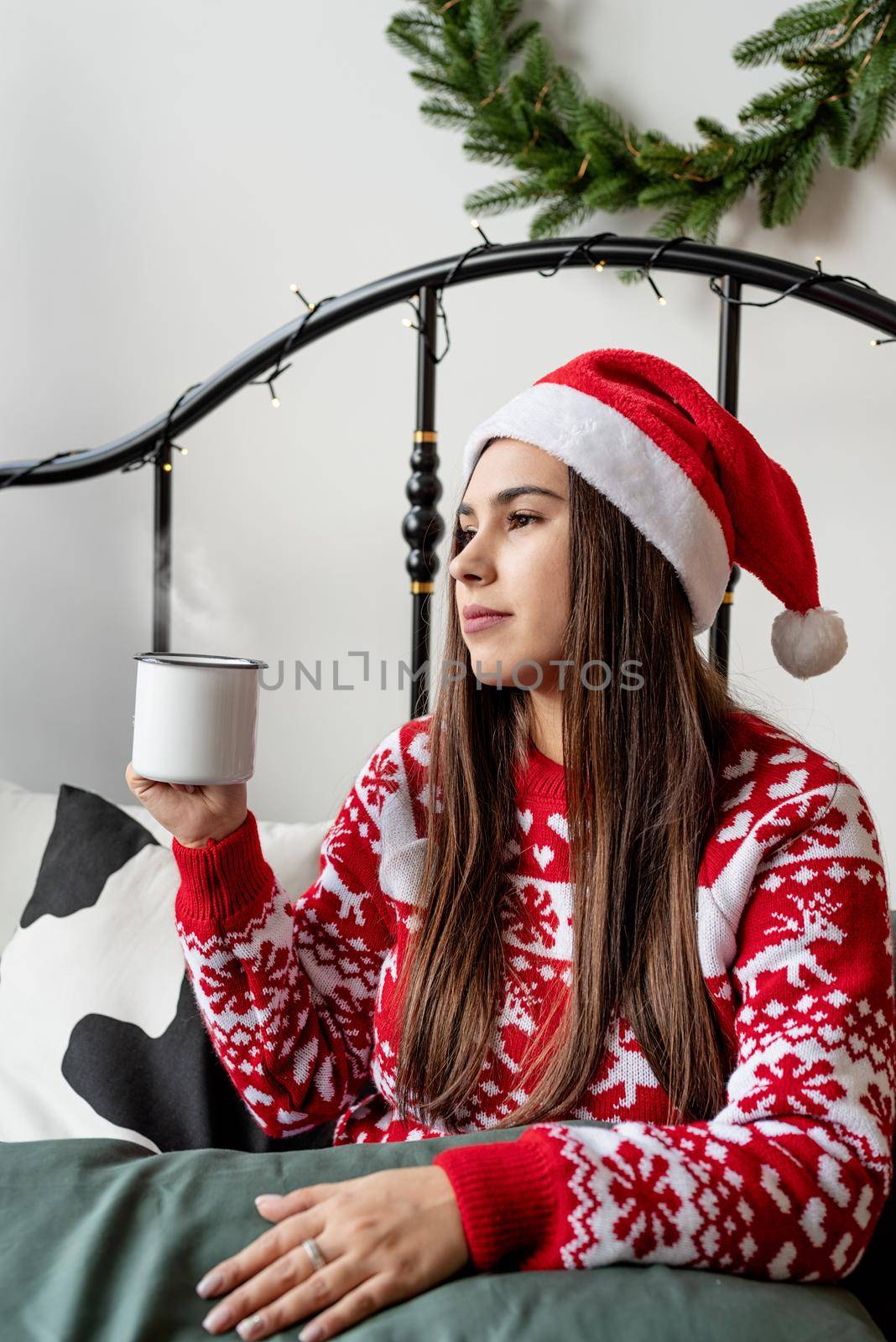 Merry Christmas and Happy New Year. Young brunette woman in red sweater and santa hat sitting on the bed celebrating christmas having cup of hot drink