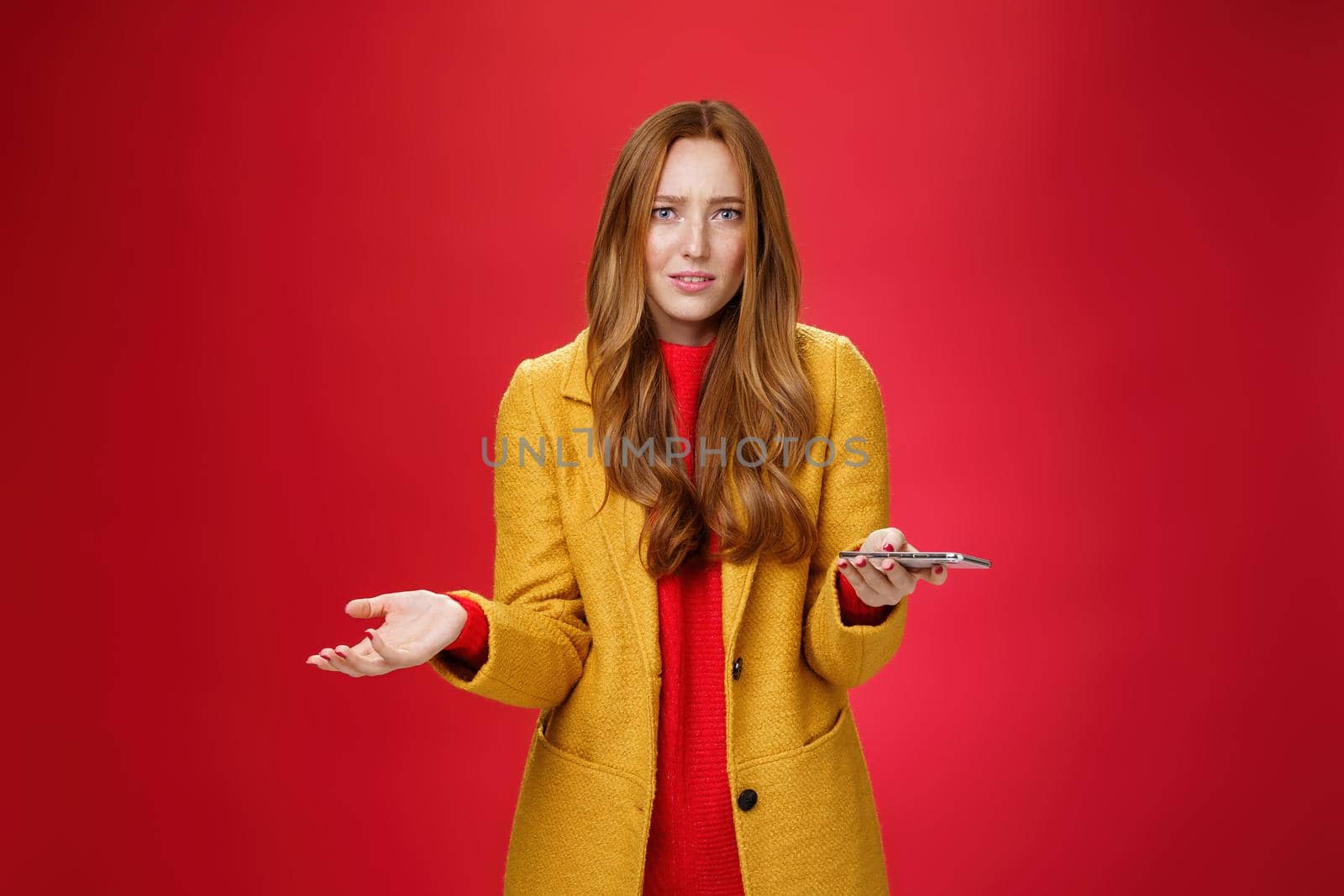 What a heck going on. Portrait of annoyed and confused perplexed ginger businesswoman in yellow coat shrugging with raised palm in clueless gesture holding smartphone troubled and questioned. Technology and dismay concept