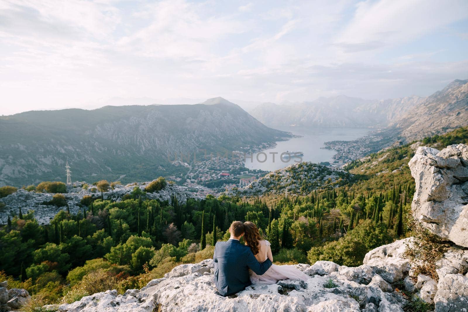 Bride and groom sit hugging on a rocky mountain and look out over the bay. High quality photo