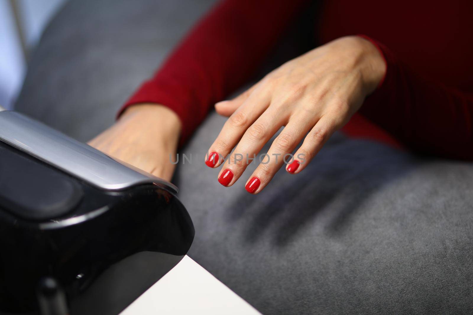 Close-up of woman client putting hand with fresh red nails under lamp to dry it. Spend time on beauty procedure in luxury studio. Beauty, wellness concept