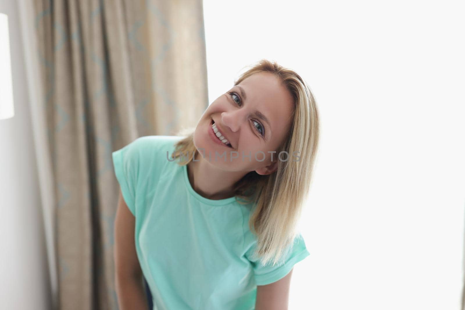 Portrait of happy smiling blonde young woman in apartment posing on curtain. Cheerful full of joy female at home. Modeling, relaxation, housewife concept
