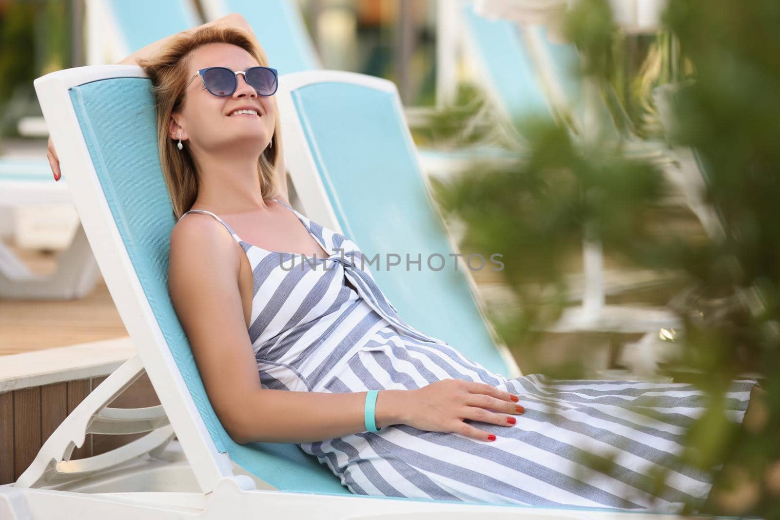 Portrait of happy young woman on sit on sunbed in pretty summer dress. Smiling relaxed female in sunglasses enjoy weather. Summer, chill, leisure concept