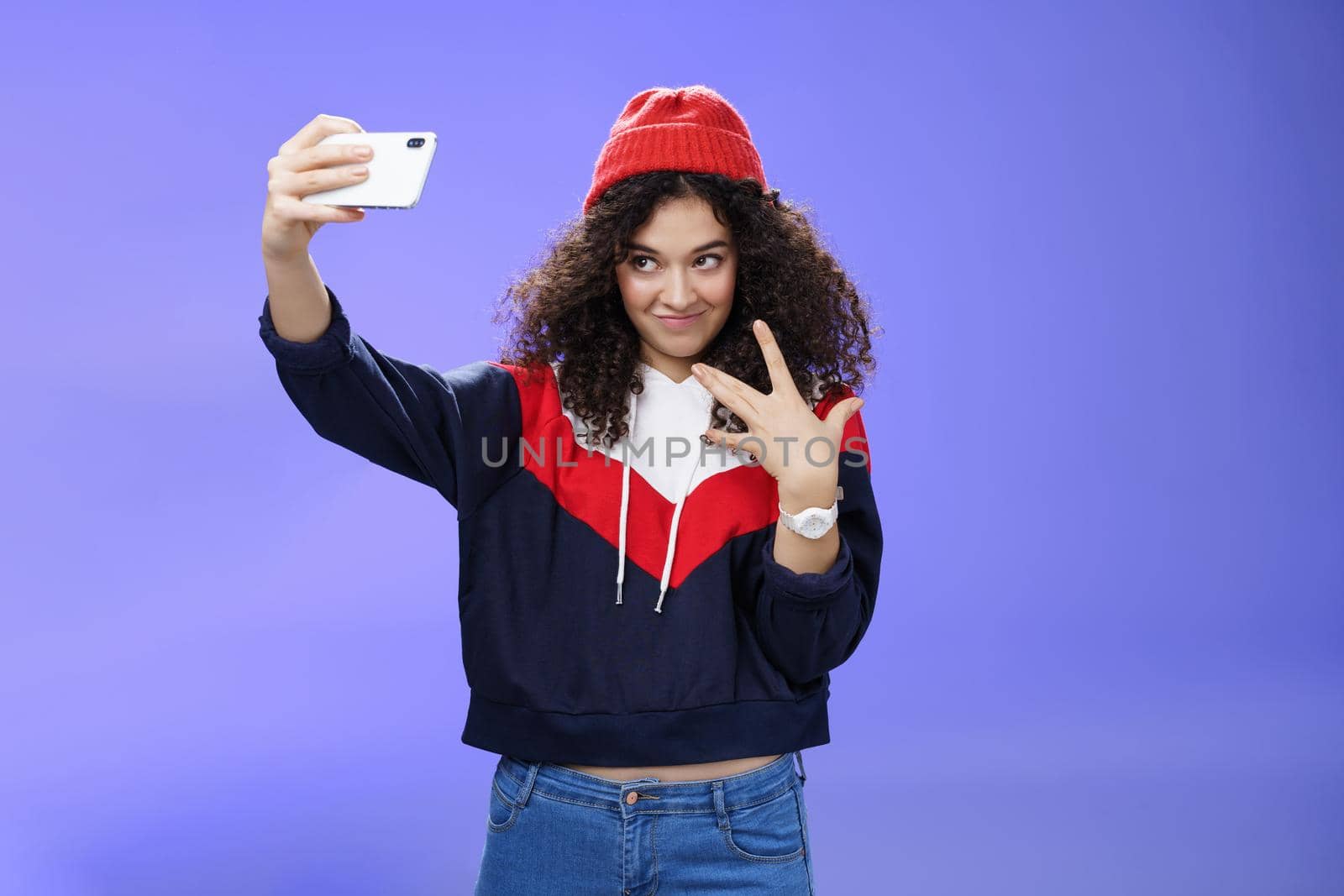 Cool and stylish good-looking caucasian female with curly hair in trendy red beanie and sweatshirt looking from under forehead awesome and swah taking selfie with smartphone holded in extended arm.