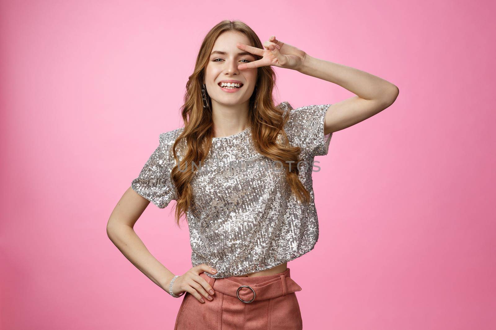 Stylish confident sassy caucasian female wearing glamour glittering blouse skirt show-off make disco peace gesture victory sign near eye smiling self-assured, posing arrogant cheeky pink background.