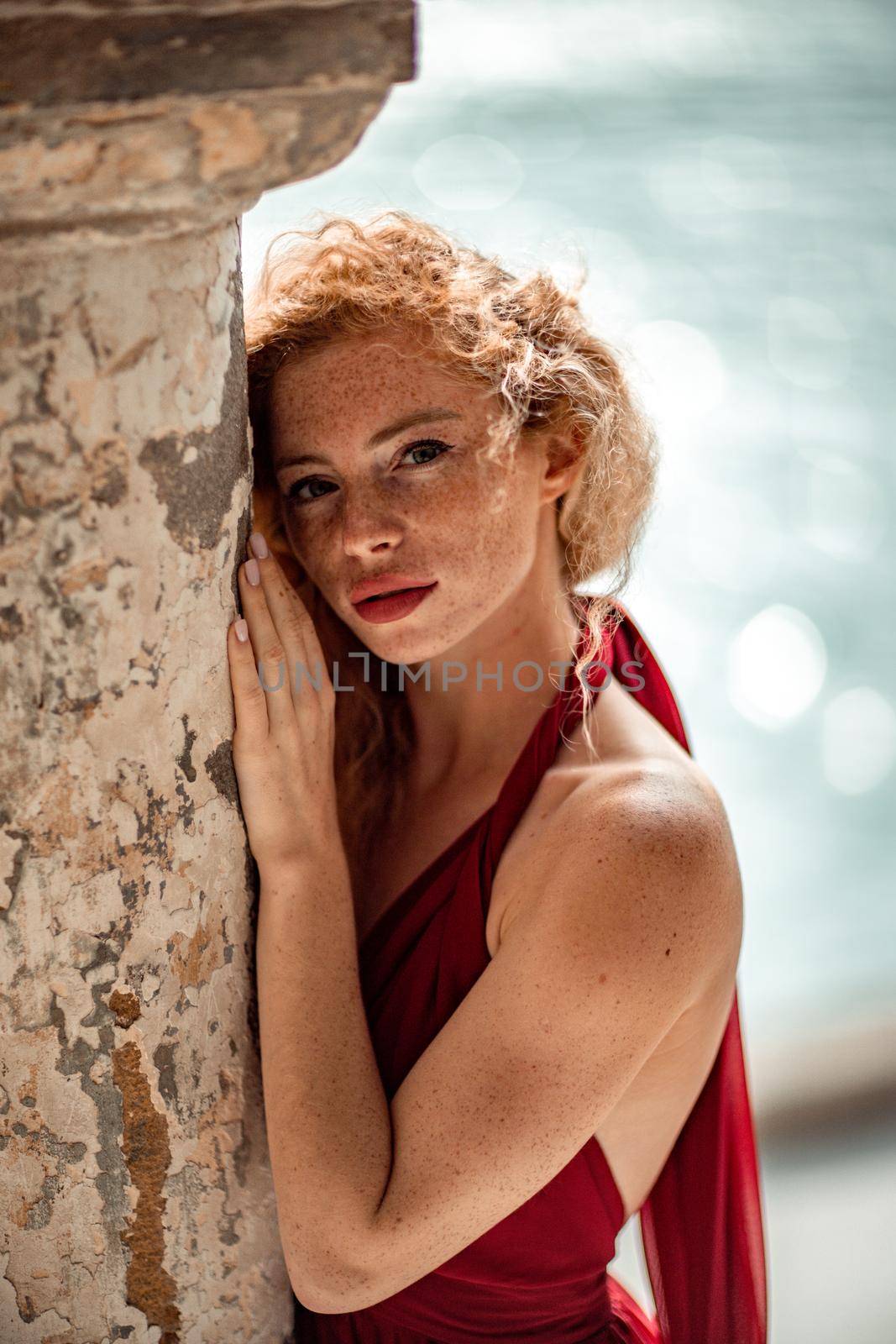 Outdoor portrait of a young beautiful natural redhead girl with freckles, long curly hair, in a red dress, posing against the background of the sea. by Matiunina