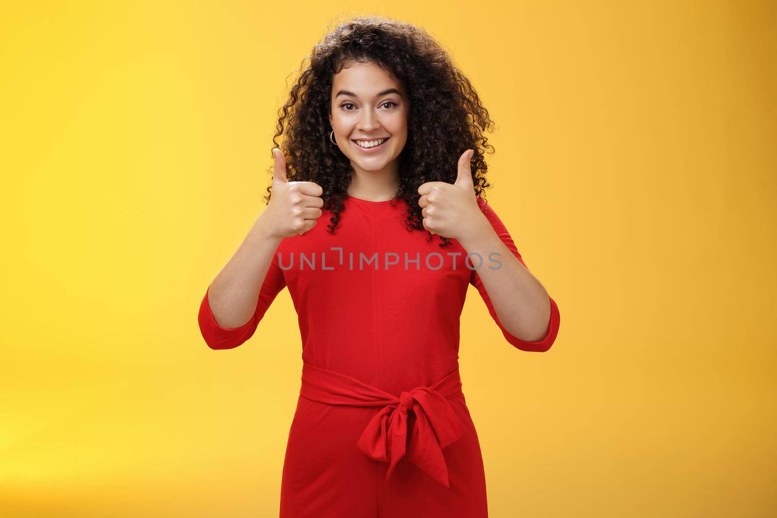 Excellent idea, nice job. Portrait of supportive delighted and happy charming female friend showing thumbs up as standing in red dress over yellow wall smiling giving positive reply, liking concept.