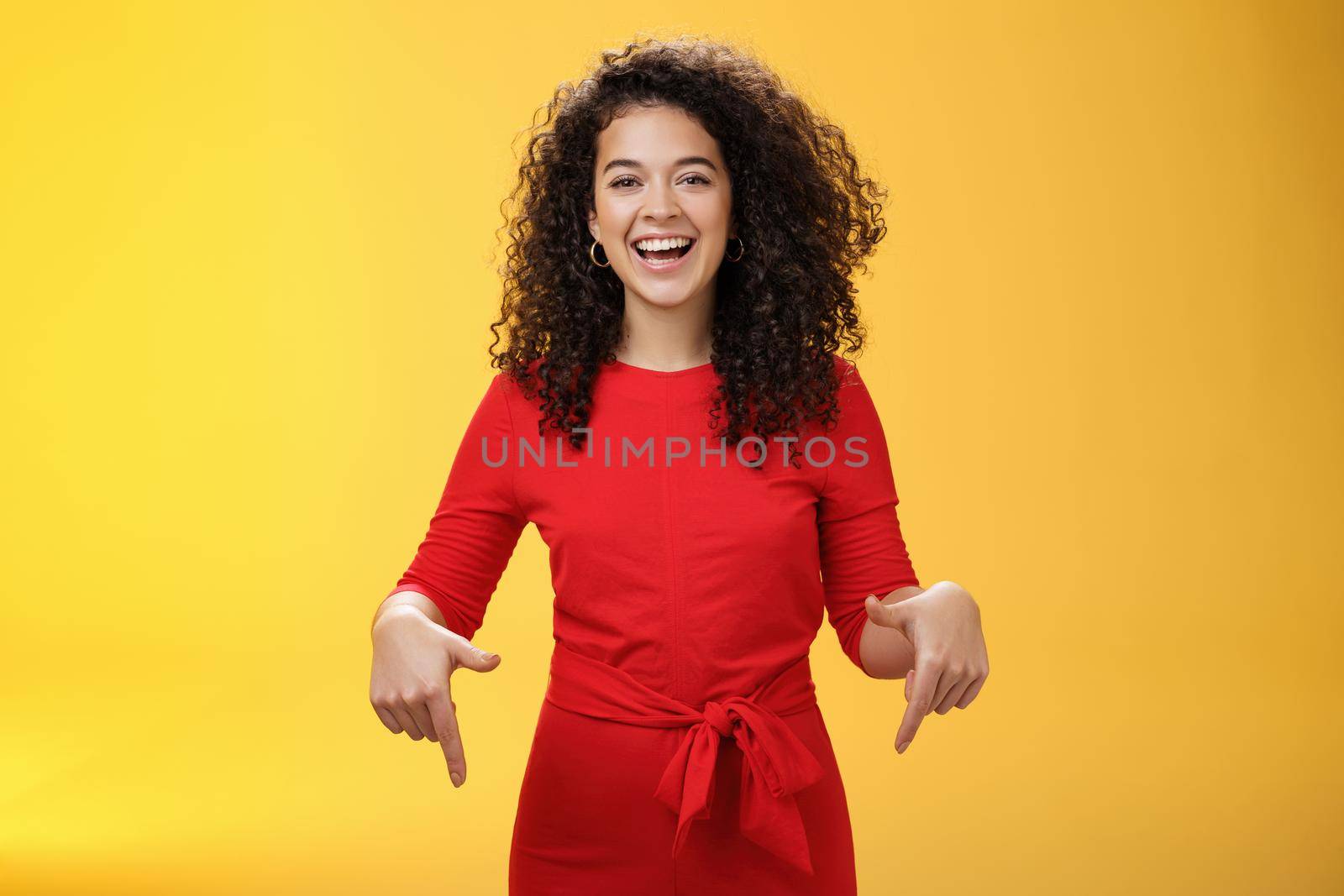 Waist-up shot of assertive charming and happy young woman with curly hairstyle laughing joyfully, smiling and pointing down as showing cool place to check out over yellow background by Benzoix