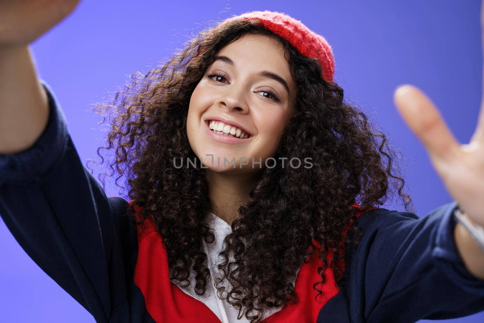 Cool and stylish friendly-looking energized happy woman in 20s with curly hair wearing warm beanie and outdoor clothes tilting head holding camera as taking selfie, smiling over blue wall.