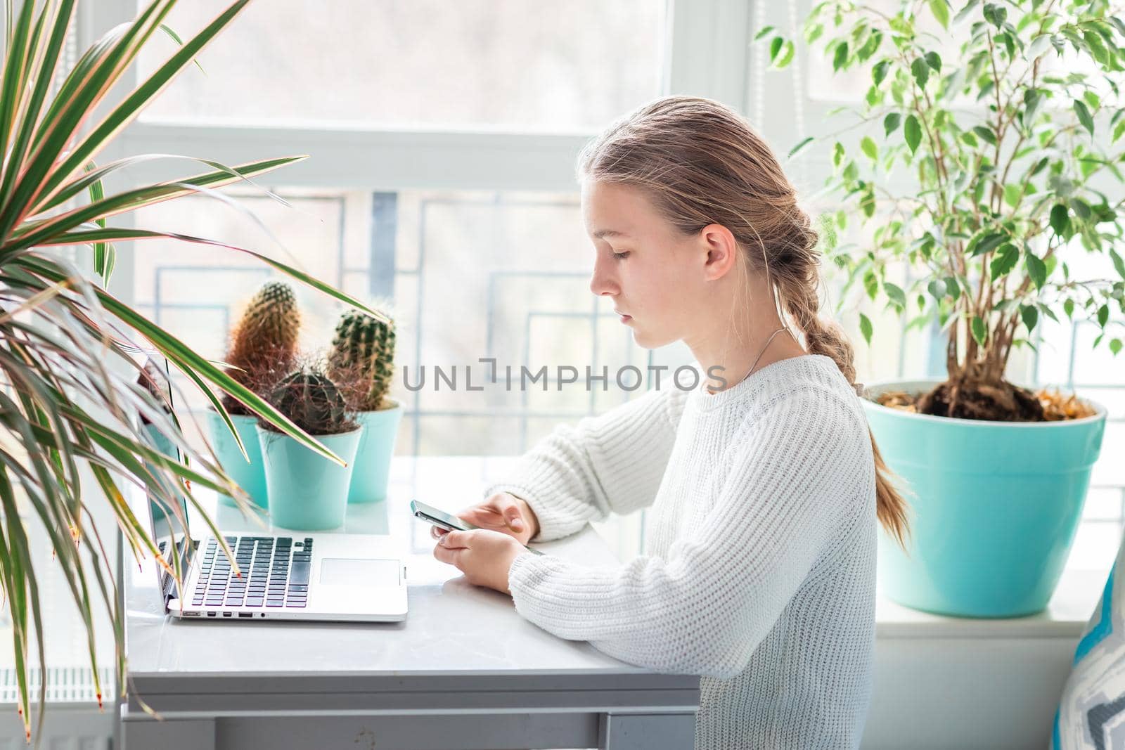 Beautiful teenage young school girl working at home checking her phone in her room with a laptop studying in a virtual class. Distance education and learning concept during quarantine