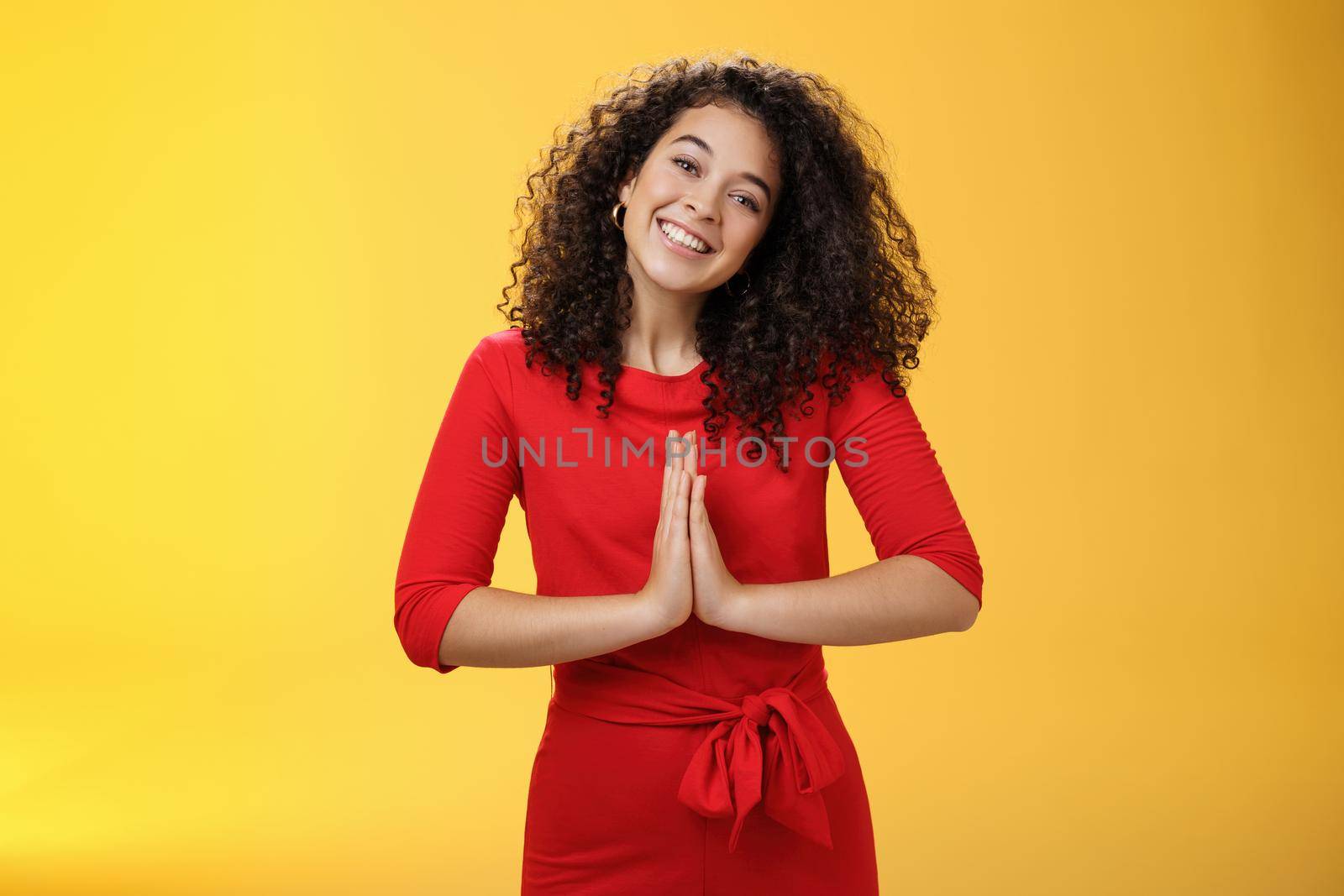 Welcome come inside. Portrait of friendly and polite good-looking female host in red dress with curly hair holding hands in namaste gesture tilting head and smiling as inviting guests in asian style.
