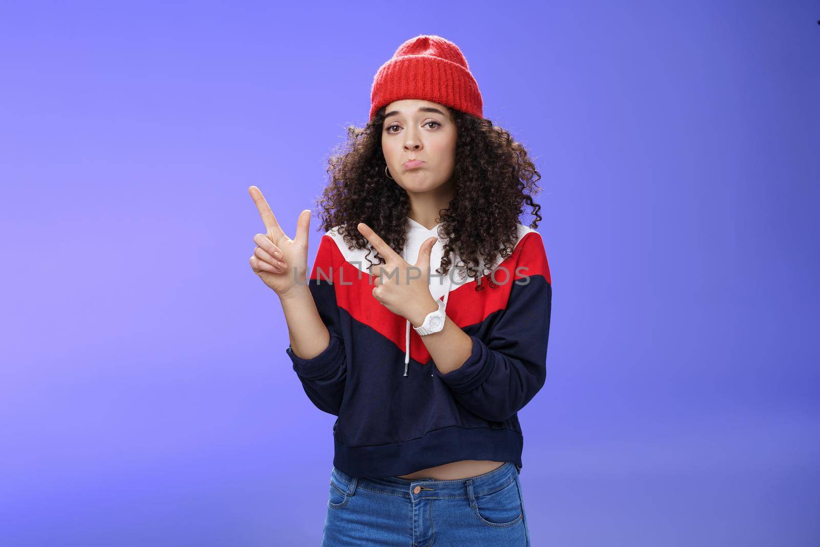 Portrait of silly upset cute curly-haired woman in trendy beanie pursing lips in sadness and frowning making disappointed, regret face pointing at upper left corner unhappy and sad over blue wall.