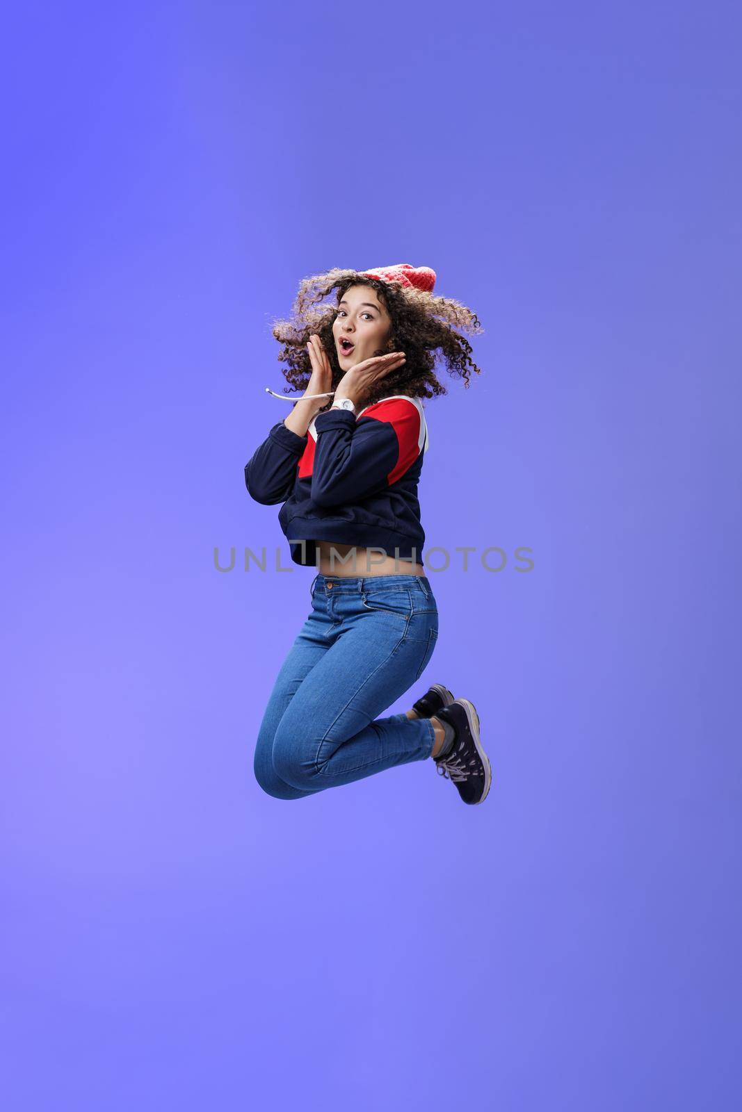 Surprised and amazed amused good-looking woman with curly hair in winter beanie and sweatshirt open mouth from joy and amazement jumping over blue background having fun feeling playful.