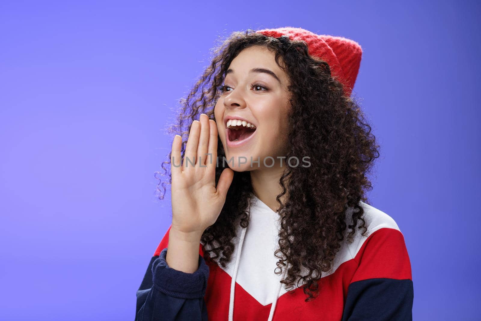 Friend shouting through street to turn attention. Portrait of carefree happy cute woman in beanie and sweatshirt holding palm near opened mouth looking with smile left as calling mate with loud yell by Benzoix