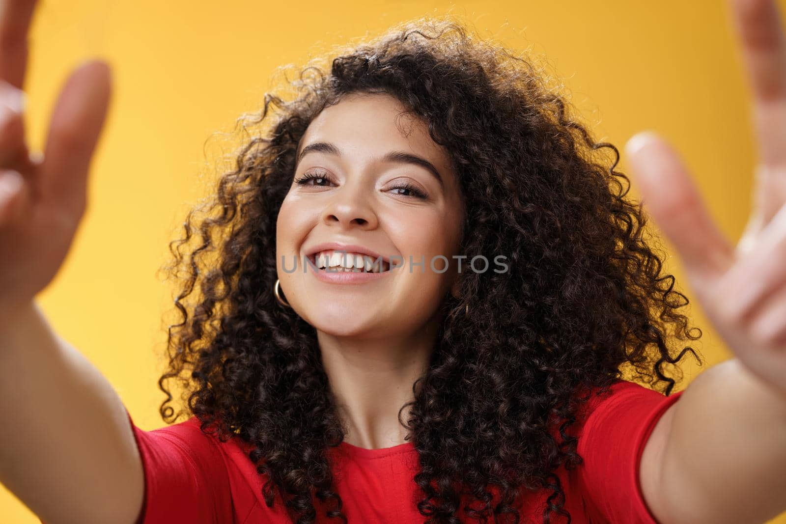 Close-up shot of friendly charming and confident young curly-haired female pulling hands towards as grabbing or hugging camera with pleased happy smile standing joyful over yellow background. Emotions and facial expressions concept