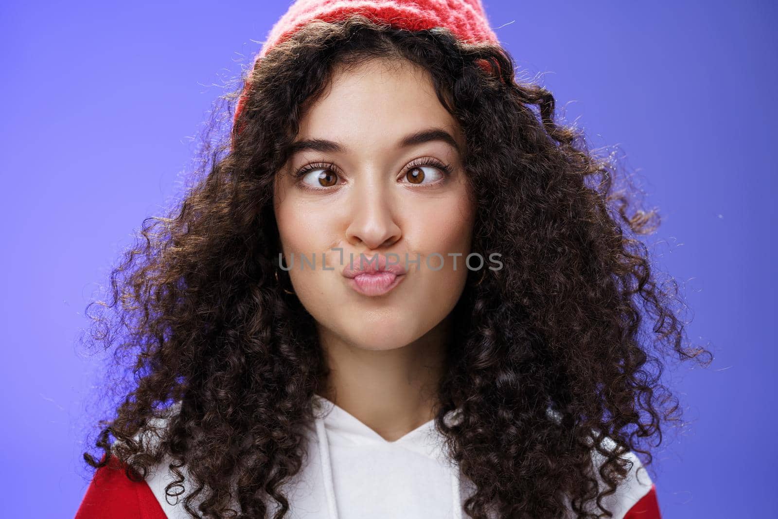 Headshot of funny and playful girl fooling around making faces not scared being funny folding lips like duck and squinting ice, aping standing childish and hilarious over blue wall in winter hat by Benzoix