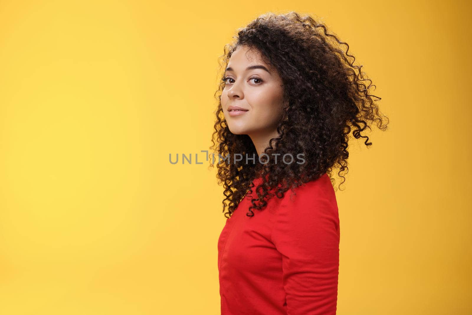 Profile shot of stylish and cool attractive curly-haired european 25s woman in red dress turning at camera and smiling broadly with delighted carefree expression, having fun over yellow background.