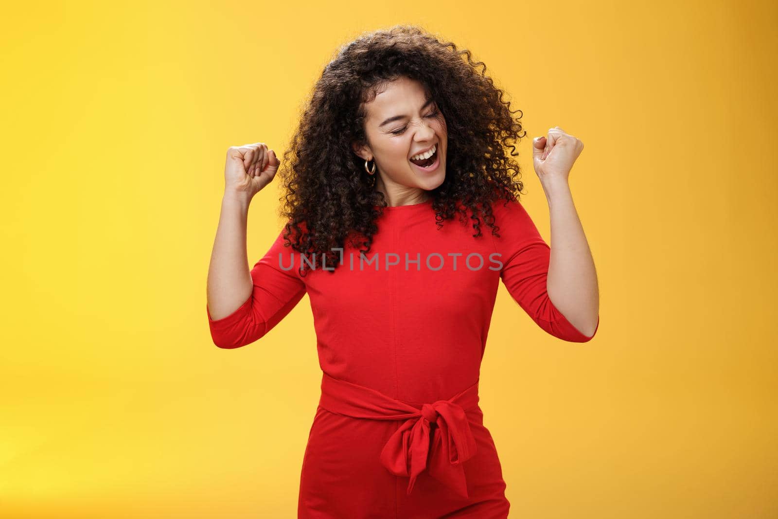 Girl enjoying cool music feeling awesome as relaxing at party dancing and feeling awesome raising clenched fists saying yeah turning head joyfully with closed eyes having fun over yellow background.