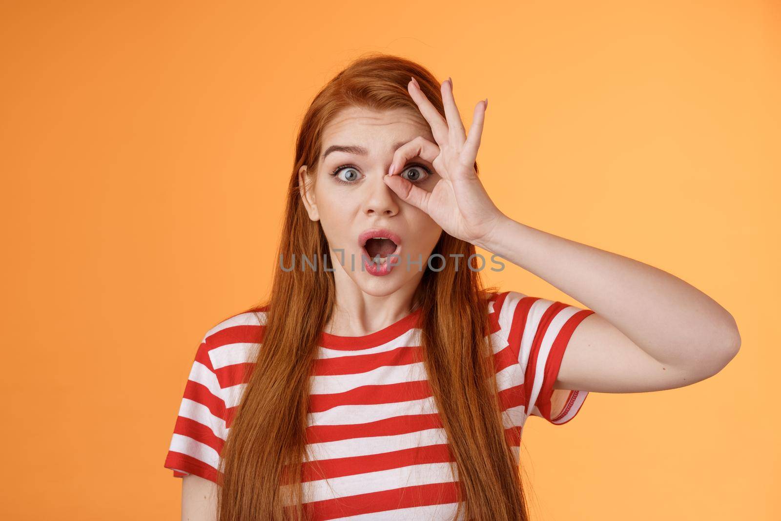 Surprised excited shocked young redhead girl gasping impressed look through okay circle, stare camera astonished awesome sale, open mouth say wow thrill and amazement, orange background.