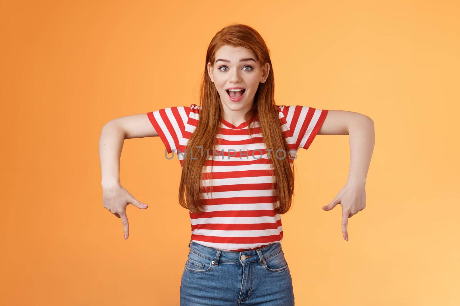 Enthusiastic lively cute redhead woman introduce incredible offer, pointing down, bottom copy space, smiling amazed, look camera, explain you awesome opportunity, feel excitement, orange background.