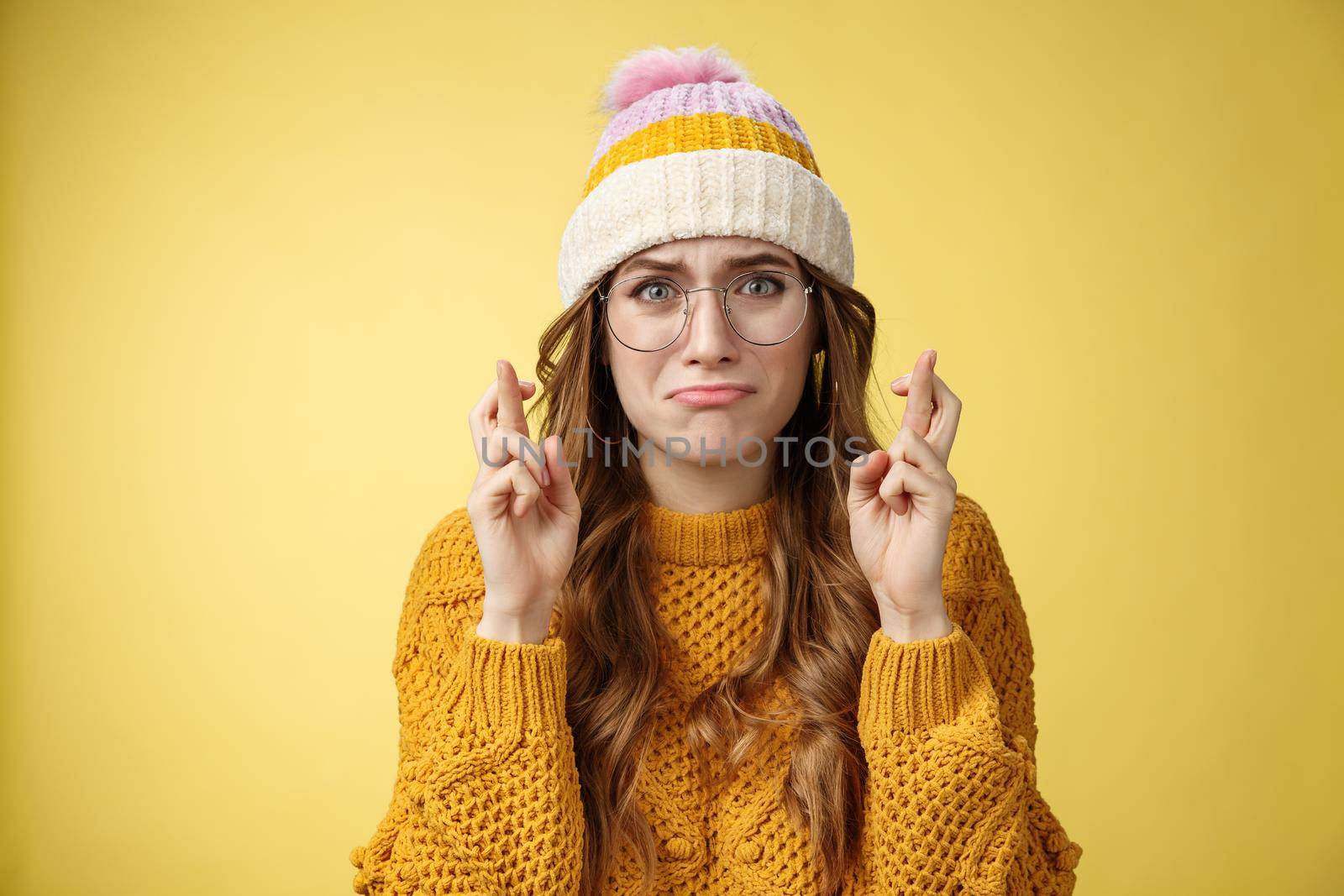 Nervous insecure unconfident cute nerd girl university student wearing glasses sulking cry, whining worried cross fingers good luck praying dream come true standing intense yellow background.