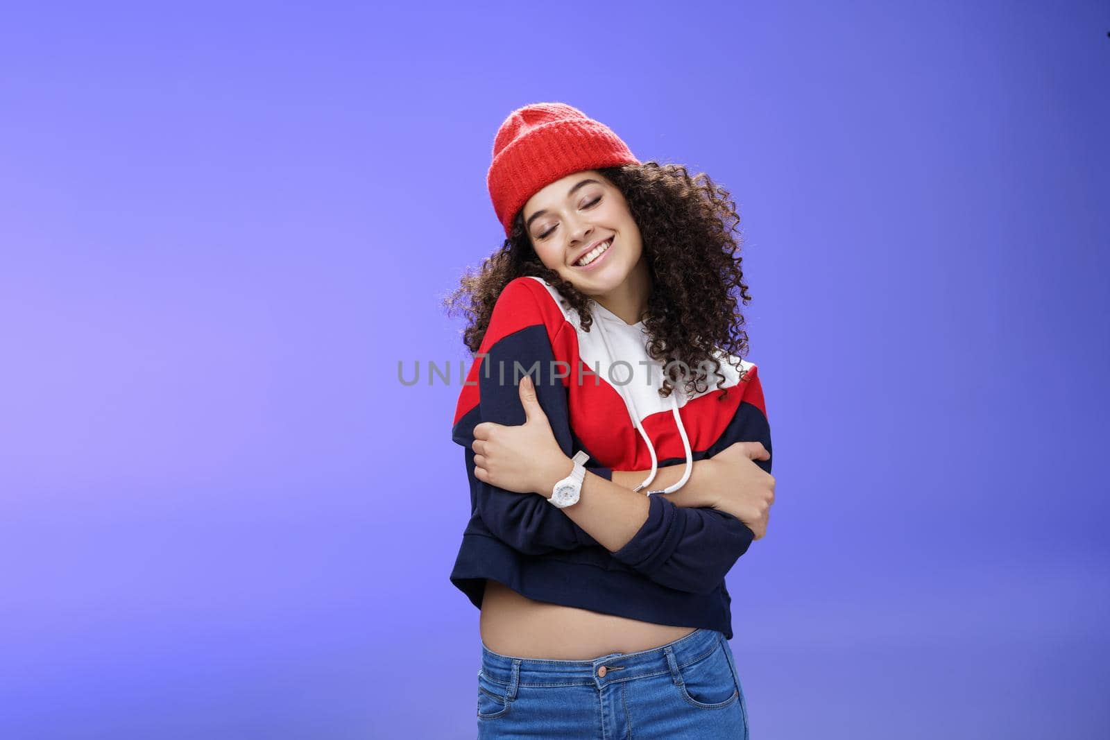 Sweet tender and sensual curly-haired woman in red stylish beanie hugging herself with closed eyes and nostalgic smile leaning on shoulder feeling warmth and tenderness as recalling loving memories.