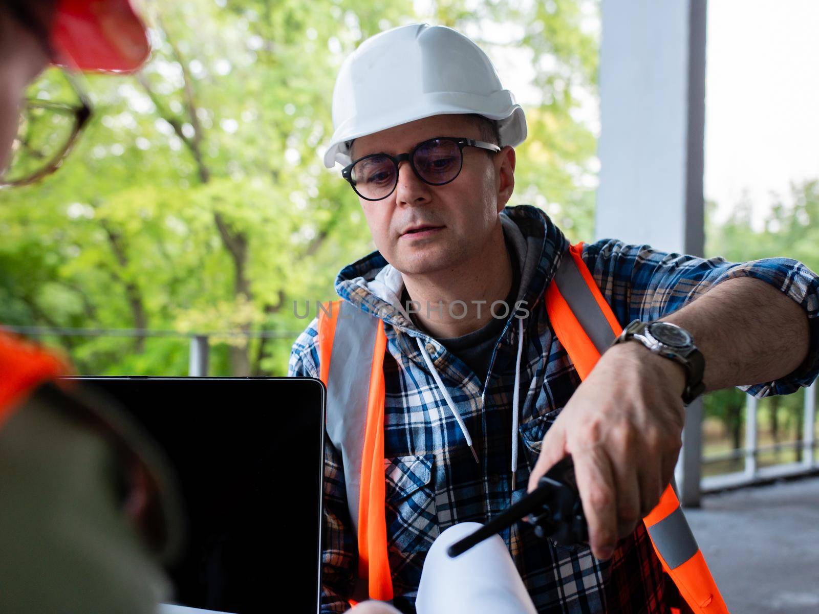 A male civil engineer at a construction site examines a building construction plan. The man is holding a walkie-talkie in his hand.