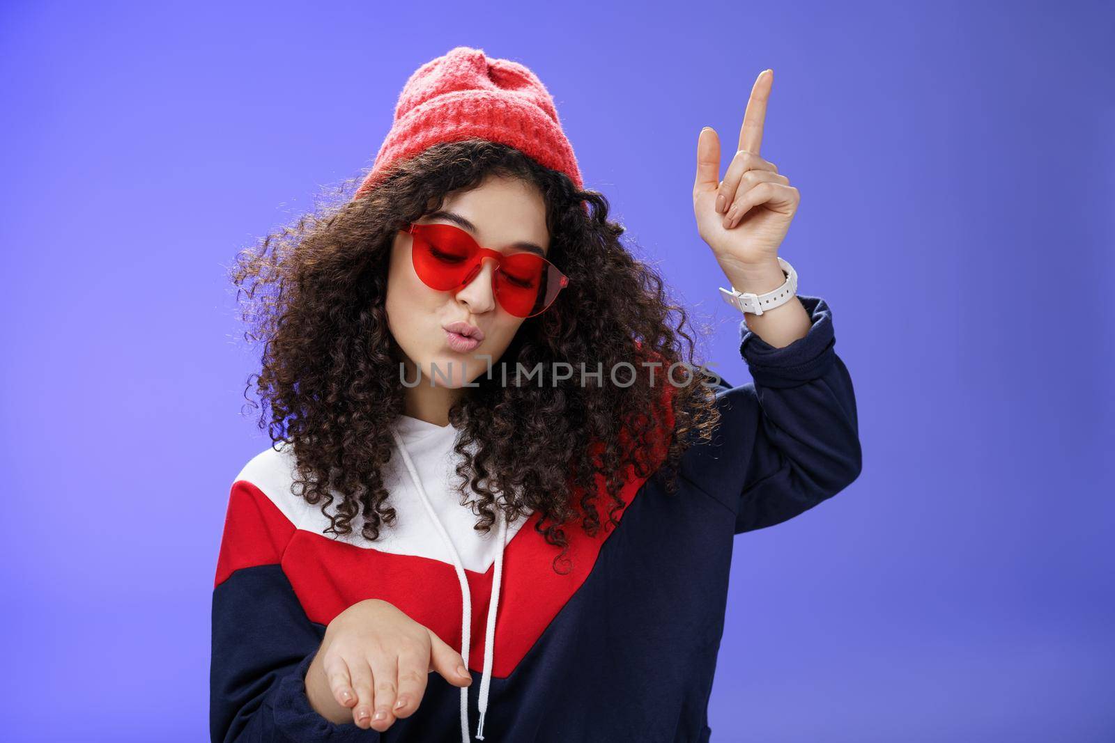 Cute party girl having fun making disco movements folding lips as enjoying cool song dancing joyfully wearing stylish red sunglasses and warm hat looking down enjoying music at party over blue wall.