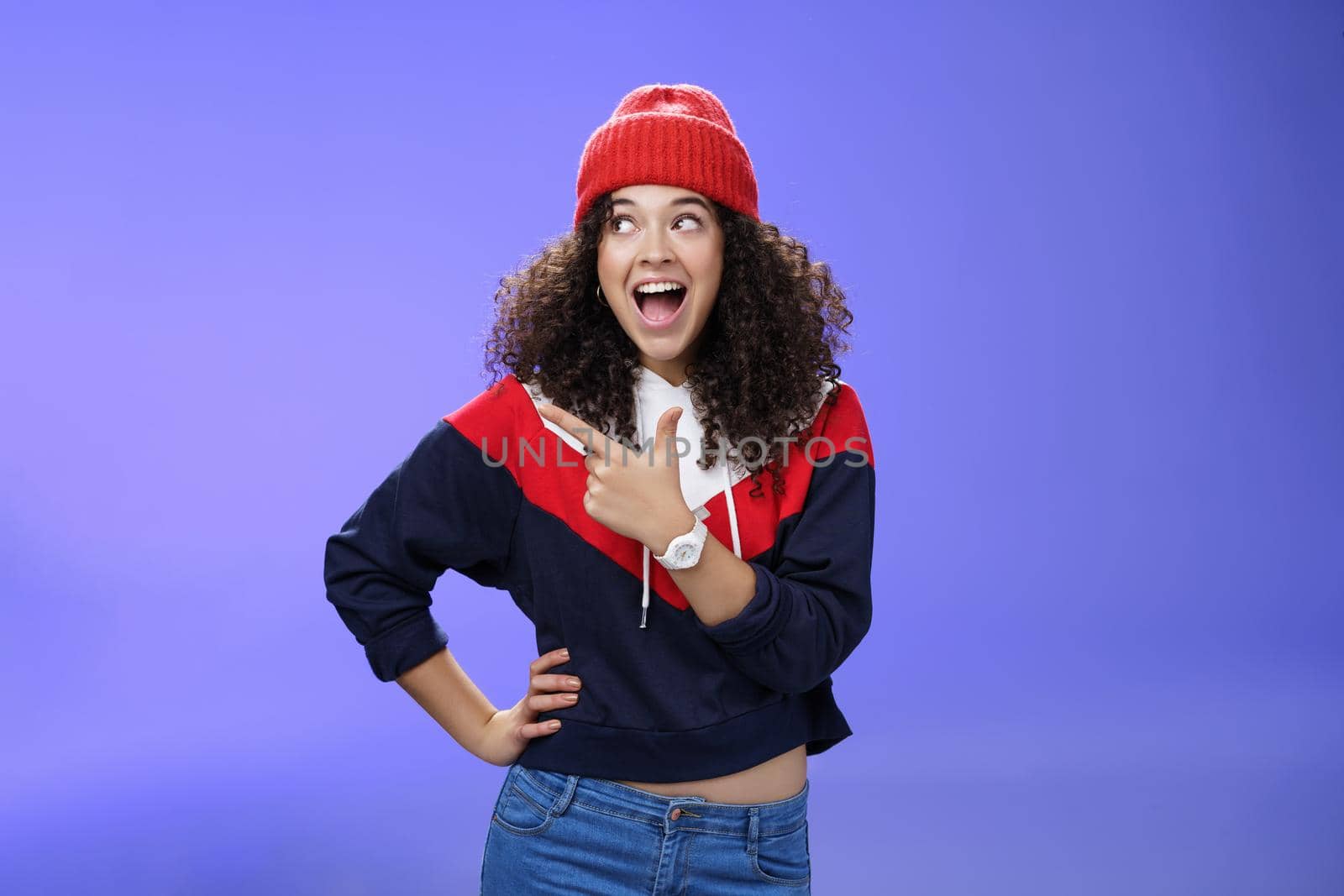 Cute and playful girl in winter stylish outfit and watch smiling broadly with excitement and joy pointing, looking at upper left corner thrilled and joyful as posing over blue background by Benzoix
